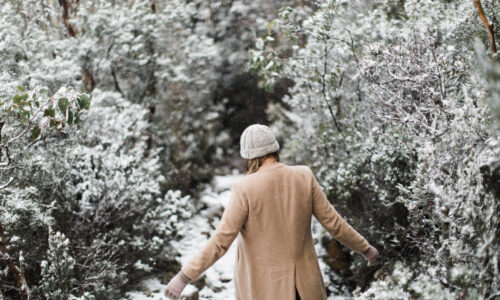 woman in wool coat and beanie walking through the fresh snow with her back to the camera. The snow has only just started to fall and the leaves on the trees are still visible through the snowflakes